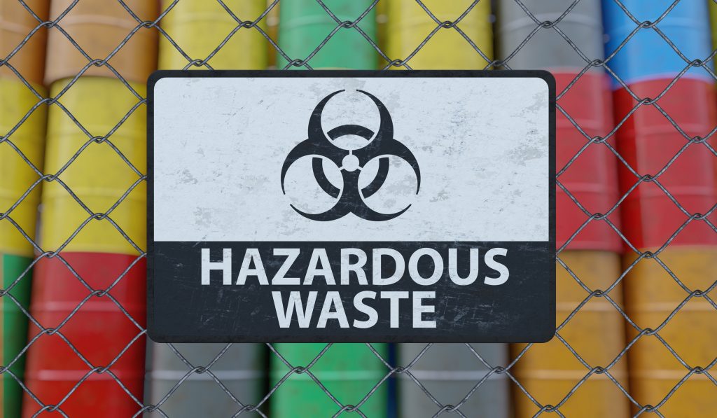 Hazardous waste sign on chain link fence. Oil barrels in background.